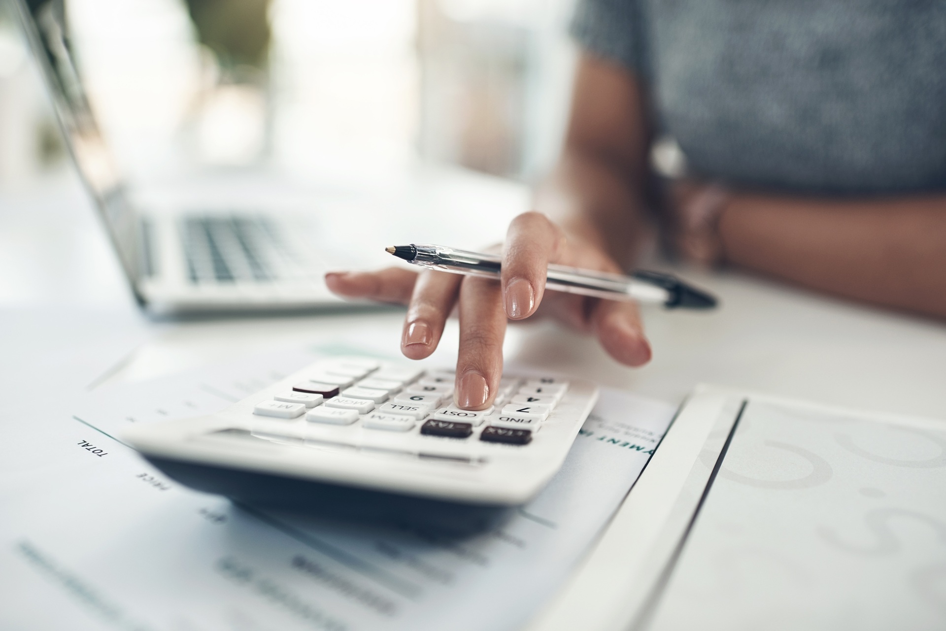 A person using a calculator while holding a pen next to an open laptop on a desk.